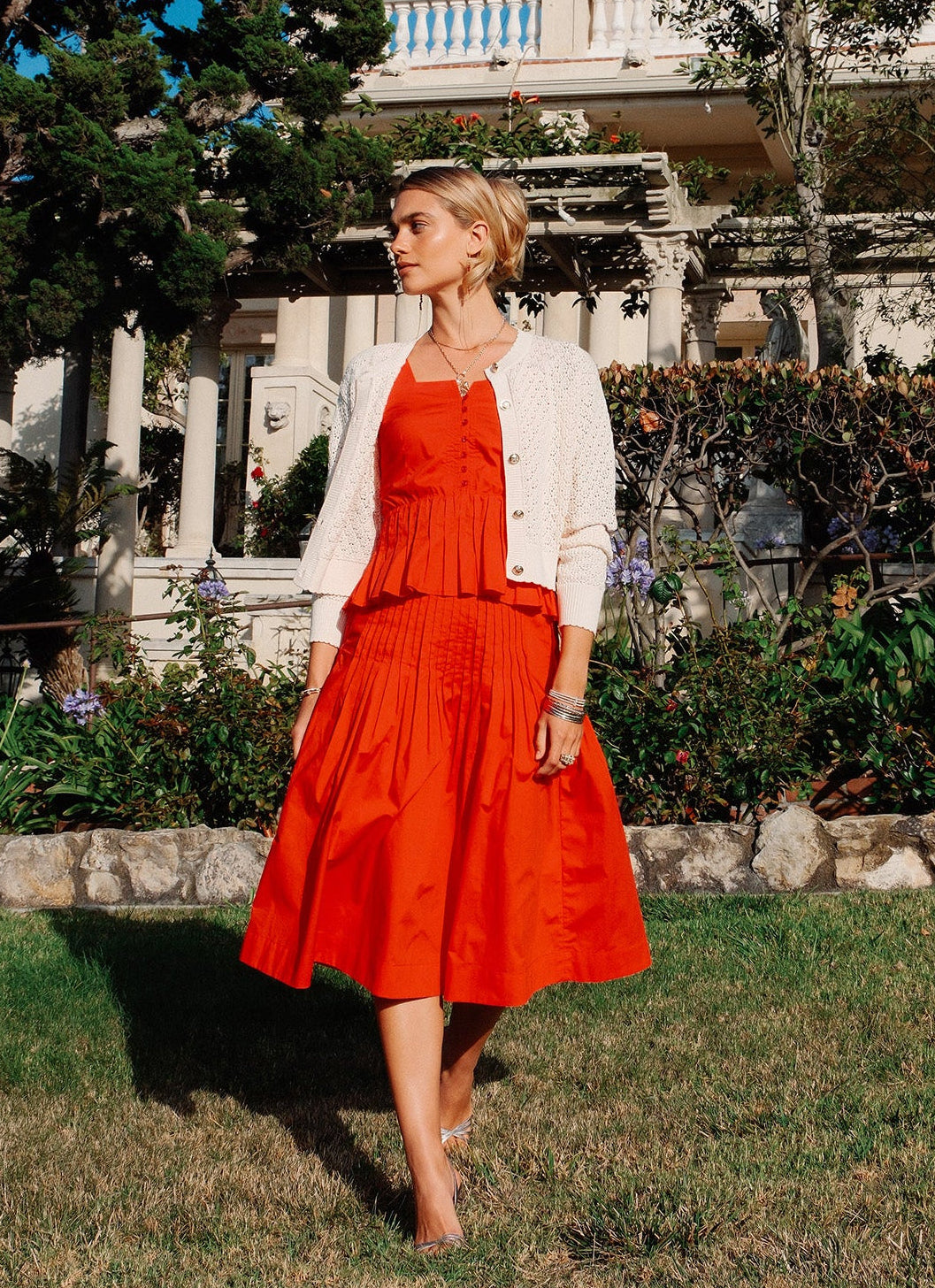 Woman in a red dress and white cardigan standing in a garden with a classical building in the background.